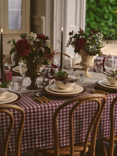 A styled dining table set with a burgundy and white gingham tablecloth, matching napkins, white plates, clear glassware, and floral centerpieces with dark red flowers and black taper candles in a warm indoor setting.