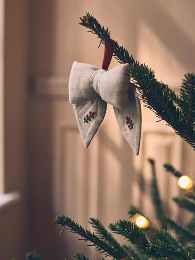 A light beige fabric bow ornament with small embroidered floral details, hanging from a green pine tree branch with warm string lights in the background.