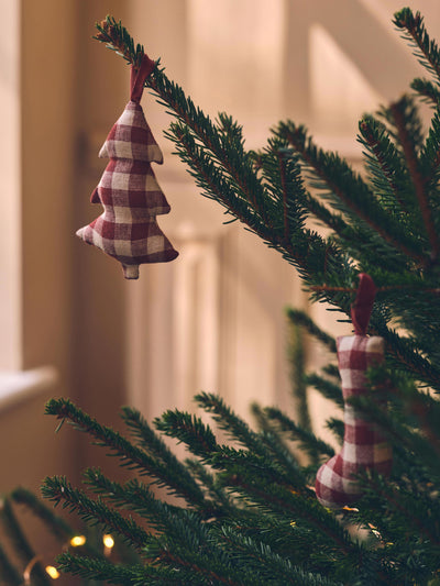 A close-up of a Christmas tree branch with two small plaid fabric ornaments hanging from the needles in warm indoor light.