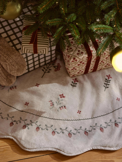 An embroidered Christmas tree skirt in cream with red and gray floral patterns, styled under a green Christmas tree with wrapped gifts and warm holiday lights.