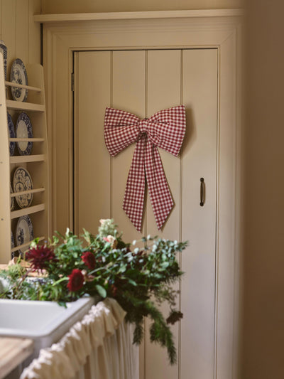 A large red and white gingham bow hangs on a cream-colored paneled cabinet door in a kitchen setting, with flowers and a dish rack visible in the foreground and background.