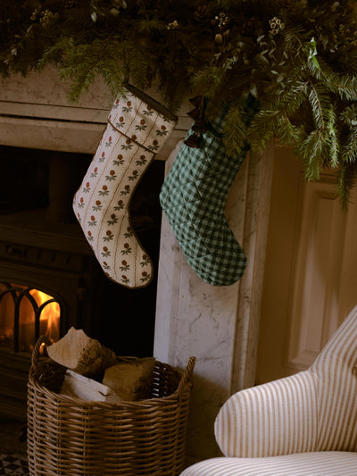 Two Christmas stockings hanging by a fireplace: one cream with a brown floral pattern and one green gingham, with pine garland, a wicker basket, and a striped armchair in a cozy indoor setting.
