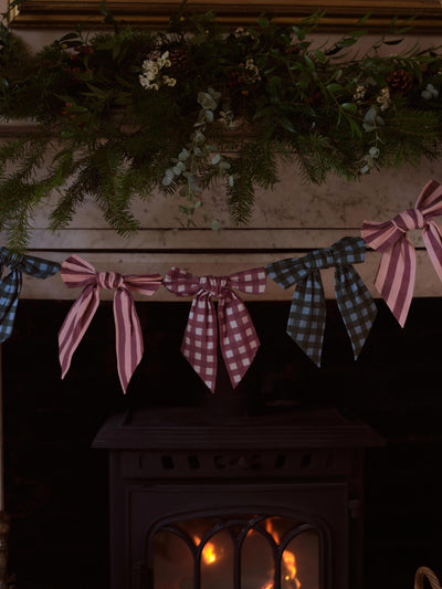 A festive garland with pink and white gingham checkered flags and pink striped flags, hung across a fireplace above a wood-burning stove. A green pine garland with white flowers decorates the mantel above.