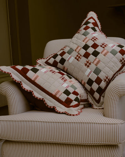Two decorative throw pillows with a rustic patchwork quilted design in brown, red, beige, and green, styled on a cream-colored striped armchair.