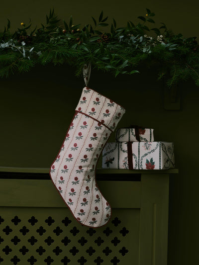 A Christmas stocking with a cream background and a red and green floral pattern hangs from a green garland above a mantel. Two wrapped gift boxes are stacked on the shelf to the right.