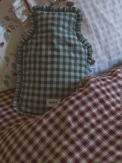 A green and blue gingham check hot water bottle with ruffled edges, resting on a bed with a matching red and white checkered blanket.