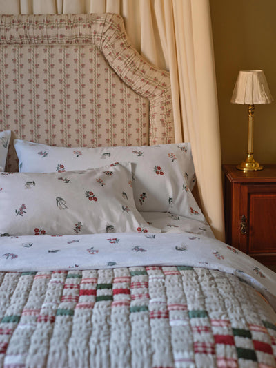 A cozy bedroom scene featuring a bed dressed in white linens with a delicate red and green floral print, a matching quilt with a patchwork design of red, green, and beige plaid and knit patterns, and a large beige floral upholstered headboard. A wooden nightstand with a gold lamp is visible on the right.
