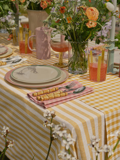 Outdoor table setting with a yellow and white checkered tablecloth, floral arrangements, and drinks.