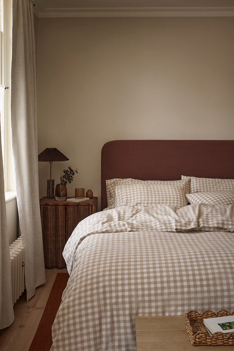 A cozy bedroom scene featuring a bed dressed in beige and white gingham check bedding with a dark reddish-brown headboard, set in a warm neutral-toned room with a wooden nightstand and lamp.