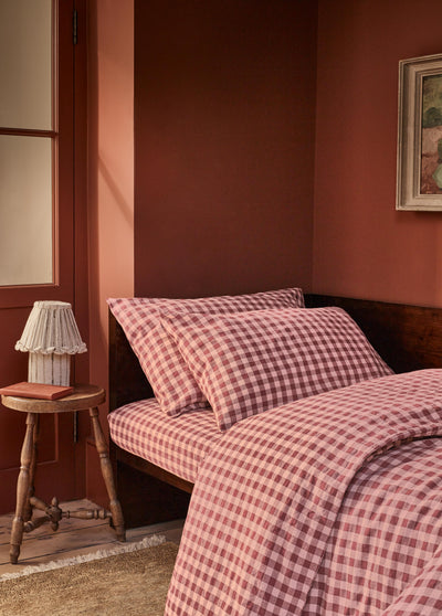 A bed dressed in a pink and red gingham check duvet cover and matching pillowcases, styled in a warm, rustic bedroom with terracotta walls, a wooden headboard, and a small wooden side table with a lamp.