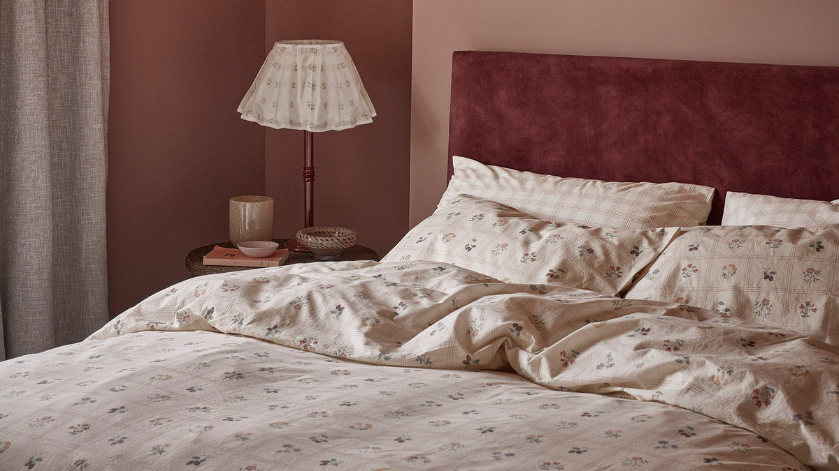 A bed dressed in a beige floral duvet cover and matching pillowcases, set against a dark burgundy velvet headboard in a warmly lit bedroom.