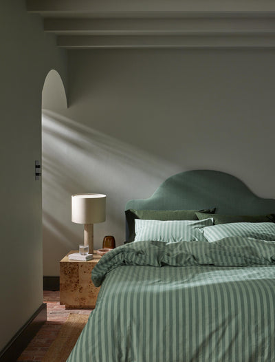 A styled bedroom scene featuring a bed dressed in sage green and white striped bedding, a matching green headboard, a wooden bedside table, and a lit table lamp in a softly lit room.