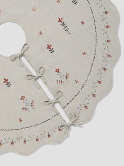 A close-up of a cream-colored fabric tree skirt with a scalloped edge, featuring delicate floral embroidery in red and gray, and three white ties with gray bows.