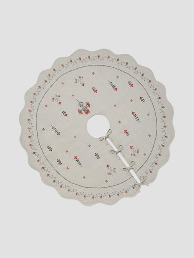 A round, scalloped-edge fabric tree skirt or doily in a light beige/cream color, decorated with a delicate red floral border and small red floral motifs, featuring a central hole and tie closures.