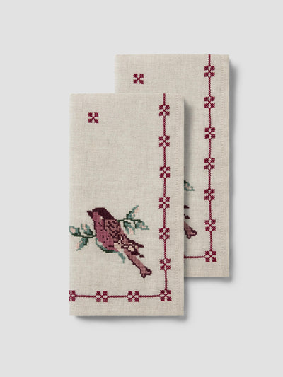 Two cream-colored linen napkins with embroidered bird and geometric border designs in burgundy and green, displayed against a light gray background.