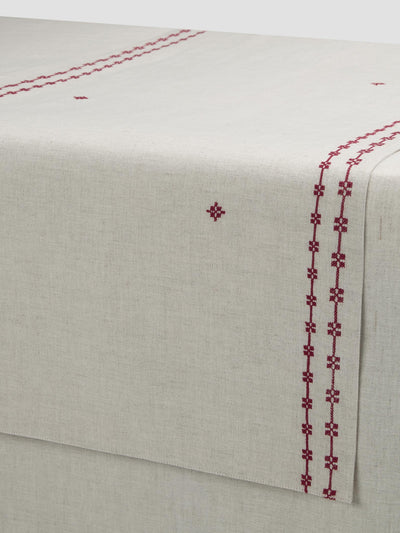 An off-white linen table runner with dark red embroidered geometric patterns along the borders and center, shown draped over a table corner.
