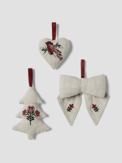 Three light beige linen Christmas ornaments hanging against a light gray background: a heart-shaped ornament with a red bird and green branch design, a tree-shaped ornament with red flowers and green stems, and a bow-shaped ornament with red floral details on each wing and a red hanging loop.