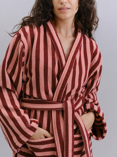 A close-up of a woman wearing a plush bathrobe with vertical stripes in dusty rose pink and deep burgundy red, featuring a matching tie belt and front pockets.