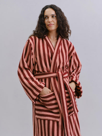 A woman wearing a plush bathrobe with vertical stripes in dark red and dusty pink, standing against a solid gray studio background.