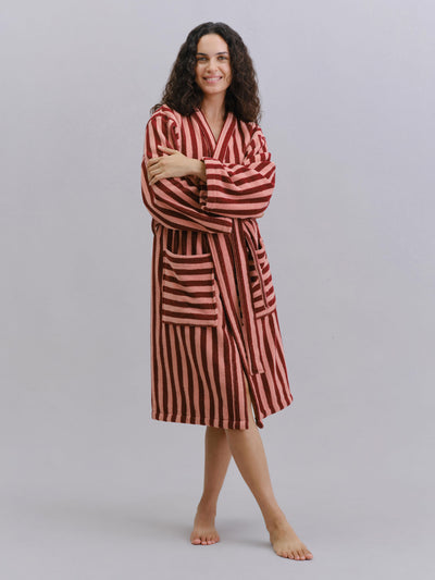 A woman modeling a long-sleeved striped bathrobe with red and dark brown vertical stripes, standing against a light gray studio background.