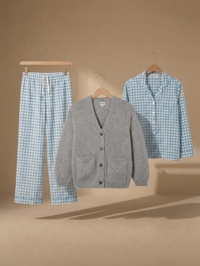 A three-piece pajama set displayed on hangers against a warm beige background. The set includes a light blue and white gingham pajama shirt, matching gingham pajama pants with a white drawstring, and a heather gray button-front cardigan.