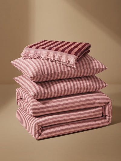 A stack of folded bedding pieces, including pillows and a folded blanket or duvet, in a dusty pink and burgundy striped pattern, set against a warm beige background.