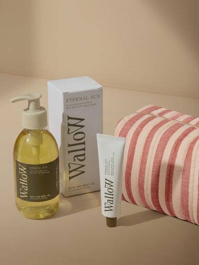 Wallow bath and body set featuring a pump bottle of Eternal Tea bath and body oil, a white box, a white tube, and a red and white striped bath towel on a beige background.