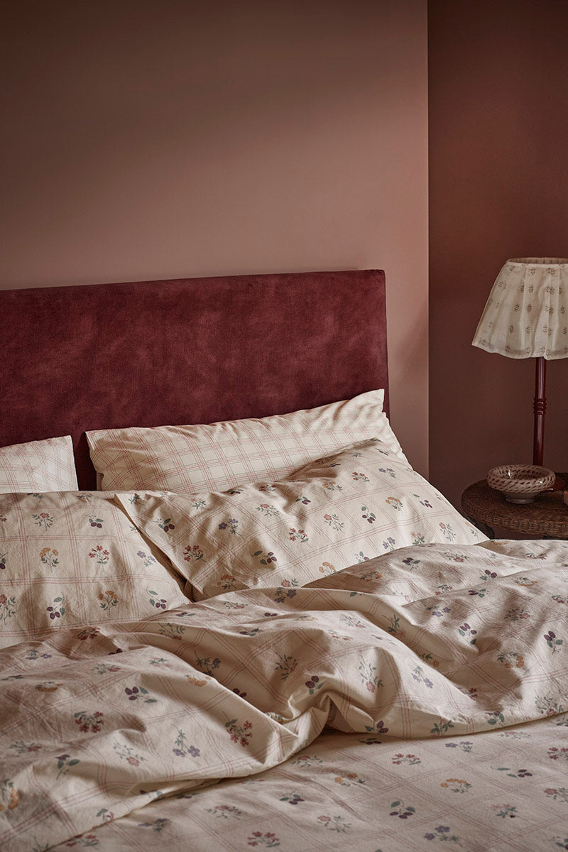 A styled bedroom scene showing a bed made with cream bedding featuring a delicate floral print, set against a deep burgundy velvet headboard and a matching wall, with a bedside lamp on a wooden nightstand.