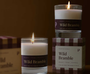 Three Wild Bramble scented candles in clear glass jars with white wax and dark purple labels, arranged on a warm brown surface with their branded boxes. One candle is lit and placed in front, another is lit and stacked on top of a box, and a third box is partially visible on the left.